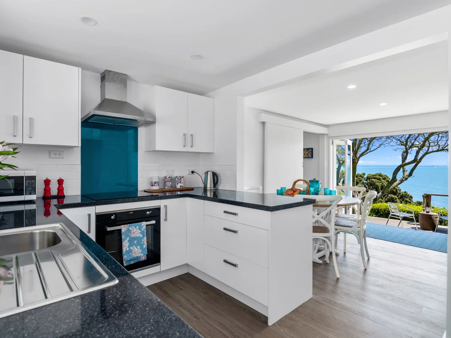 A bright kitchen opens onto a dining area beside large windows overlooking the ocean.