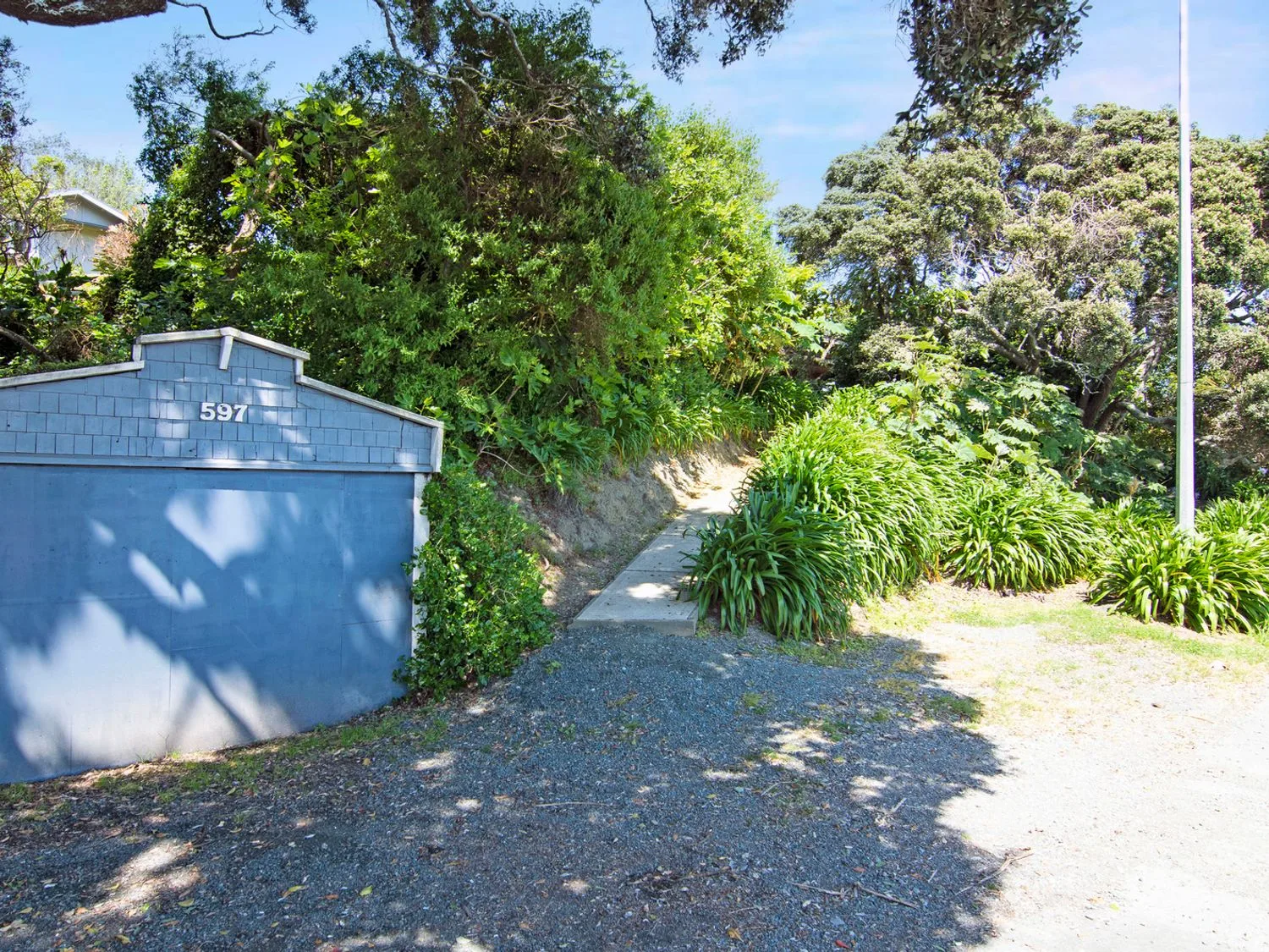 Un acceso de grava que conduce a un garaje azul con el número 597 y un sendero de hormigón junto a arbustos verdes y árboles en Bayview Bungalow - Nelson Holiday Home, en Nelson.