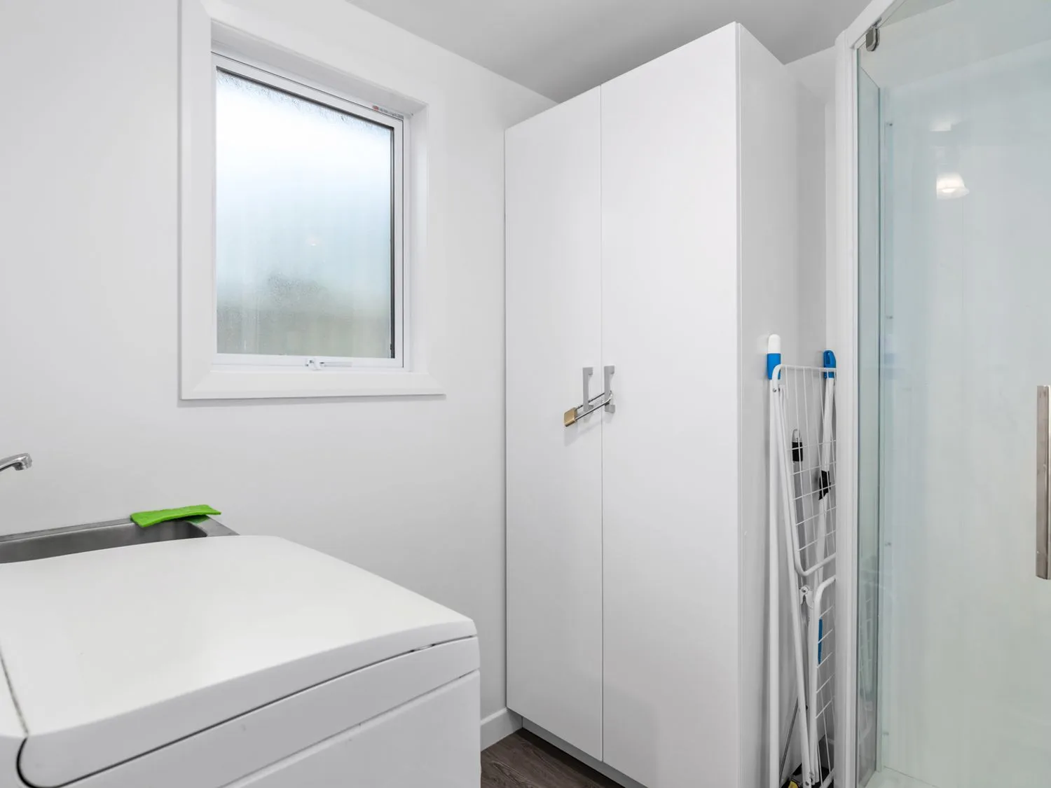 Un cuarto de lavado con una lavadora, un lavadero, una ventana, un armario blanco y un tendedero plegado apoyado contra el armario en Bayview Bungalow - Nelson Holiday Home, en Nelson.
