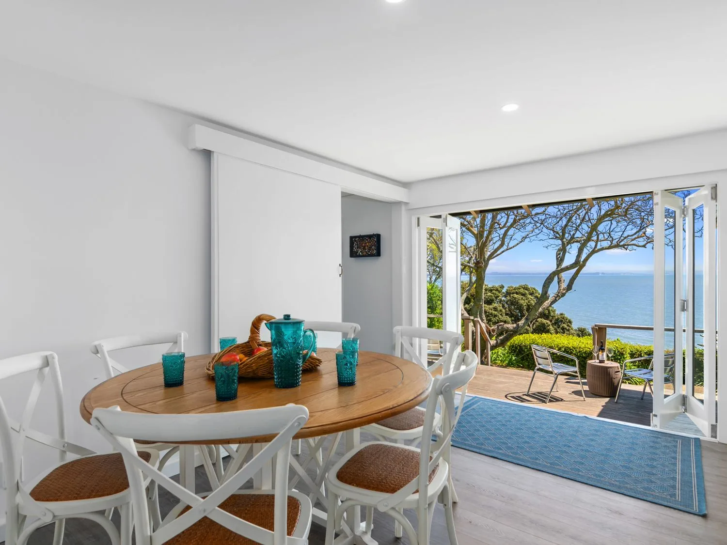 A dining area with a round wooden table and white chairs next to an open door leading to a deck with chairs and an ocean view at Bayview Bungalow - Nelson
