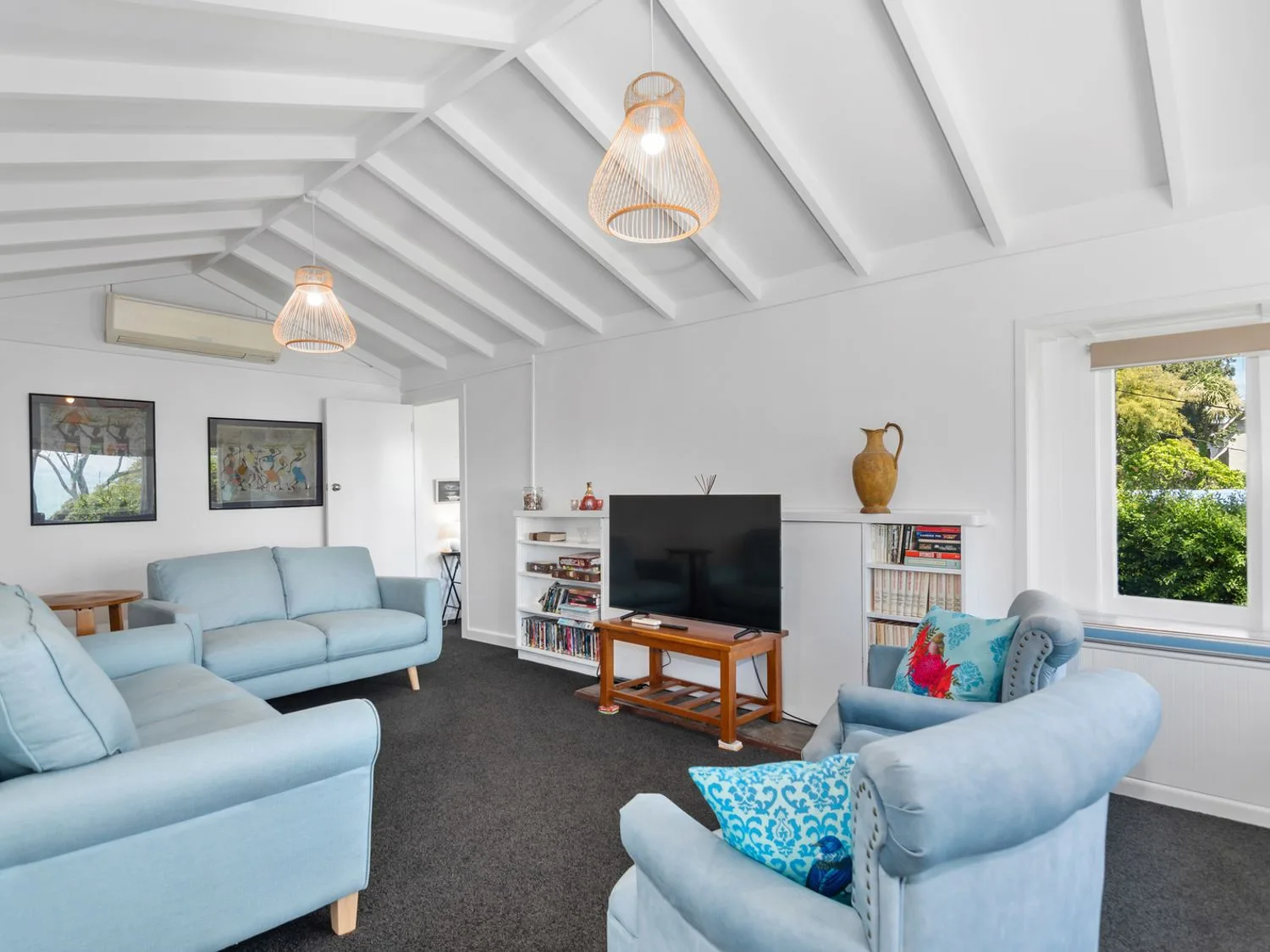 Una sala de estar con sofás azules, un televisor sobre un mueble de madera, estanterías para libros y cuadros enmarcados en la pared en Bayview Bungalow - Nelson Holiday Home, en Nelson.