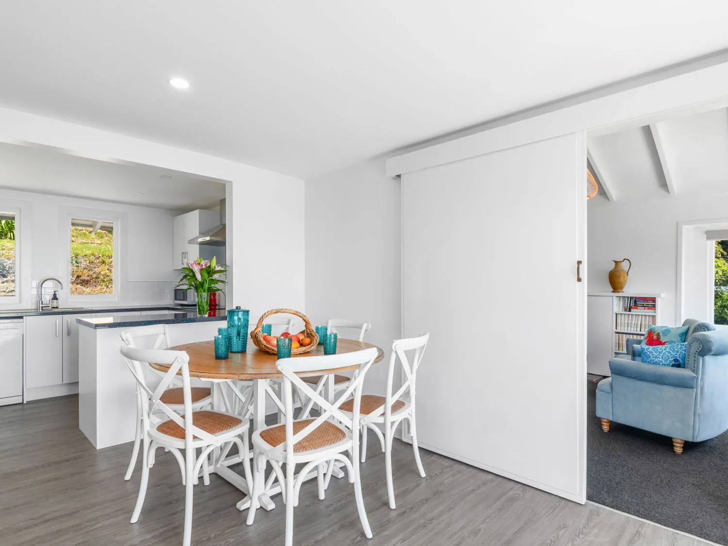 A dining area with a round wooden table and six white chairs next to a kitchen with white cabinets and a sliding door leading to a living room with a blue armchair at Bayview Bungalow - Nelson Holiday Home in Nelson