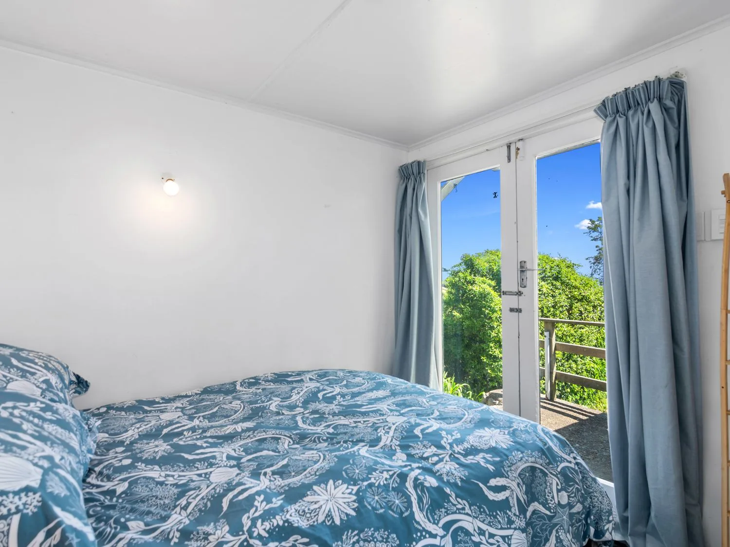 Un dormitorio con una cama con ropa de cama estampada en azul y blanco junto a puertas de cristal con cortinas azules en Bayview Bungalow - Nelson Holiday Home, en Nelson.