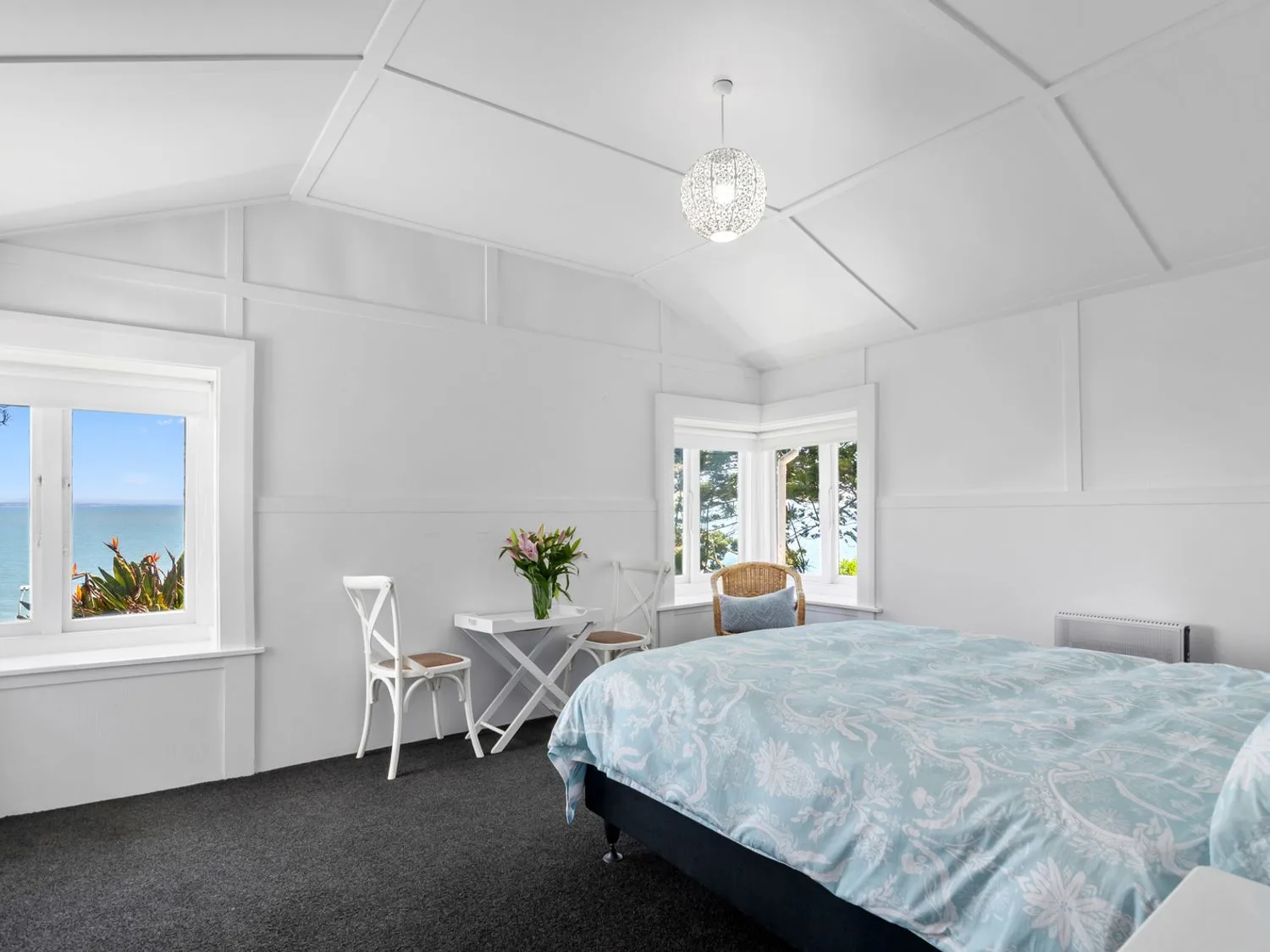 Un dormitorio con una cama, una mesa pequeña con dos sillas, una silla de mimbre y ventanas con vistas al mar en Bayview Bungalow - Nelson Holiday Home, en Nelson.