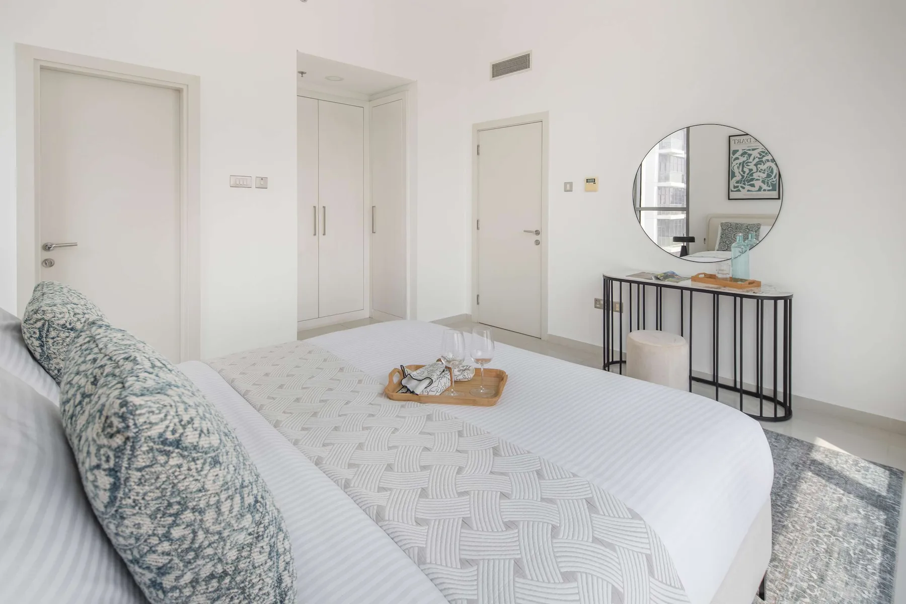 Un dormitorio luminoso con una cama bien tendida y una consola bajo un espejo redondo.
