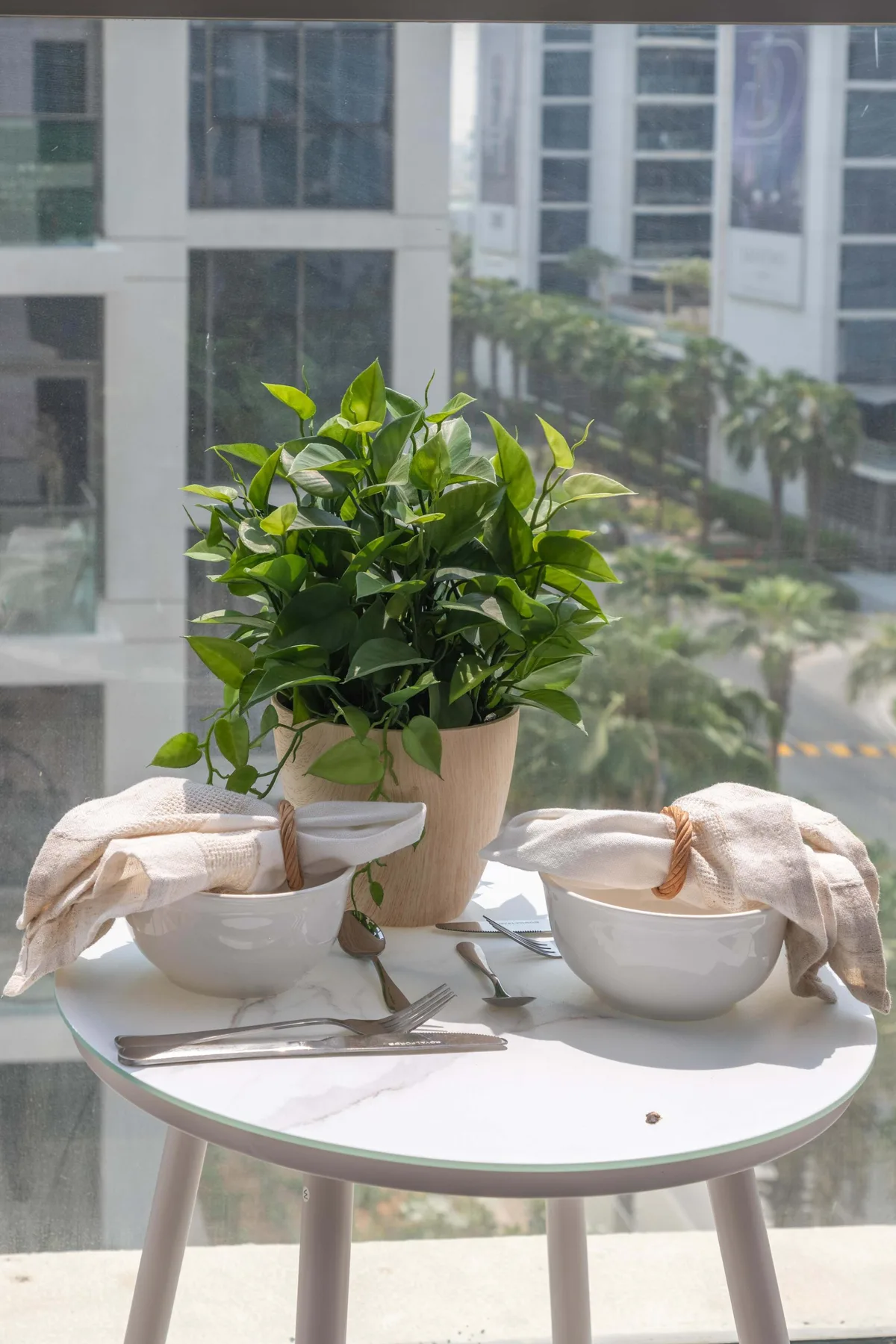 Una mesa pequeña con cuencos, cubiertos y una planta frondosa junto a grandes ventanales.