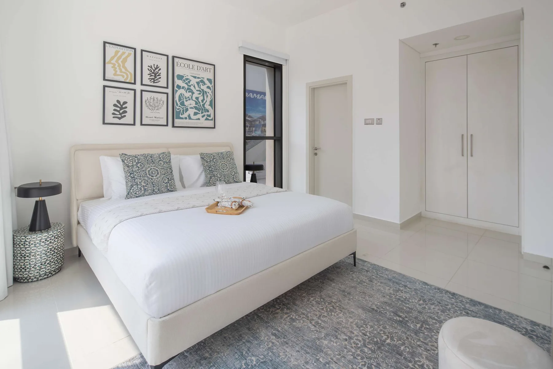 Un dormitorio luminoso muestra una cama bien tendida junto a una ventana alta.