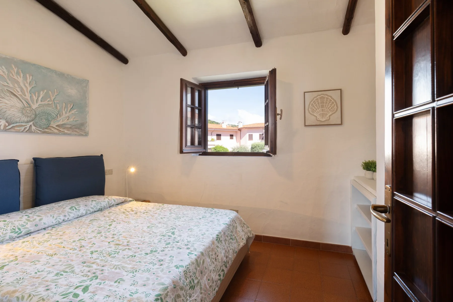 A bedroom with a patterned bedspread, dark ceiling beams, and an open window.