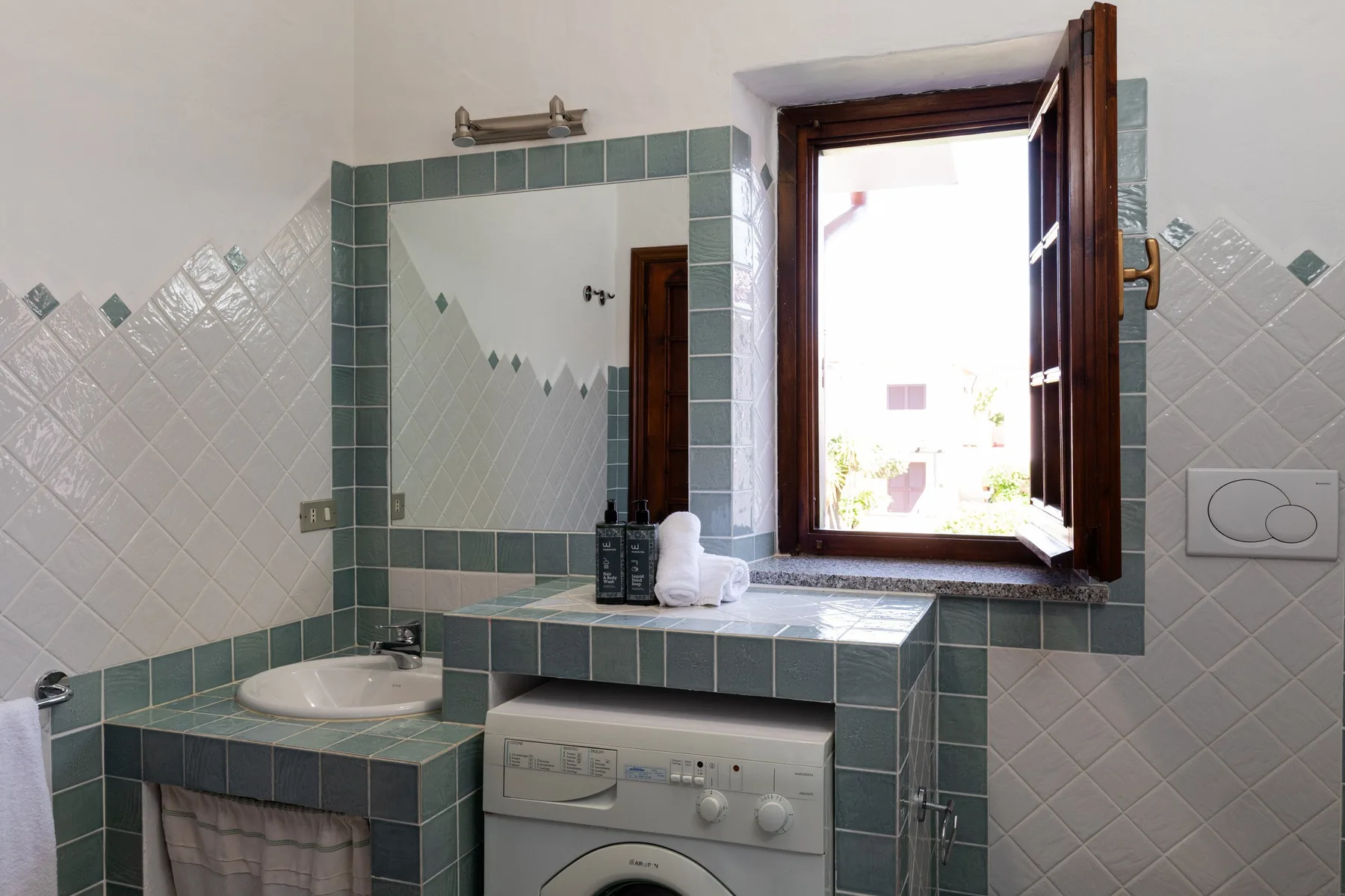 A tiled bathroom vanity with a sink, mirror, and washing machine beneath an open window.