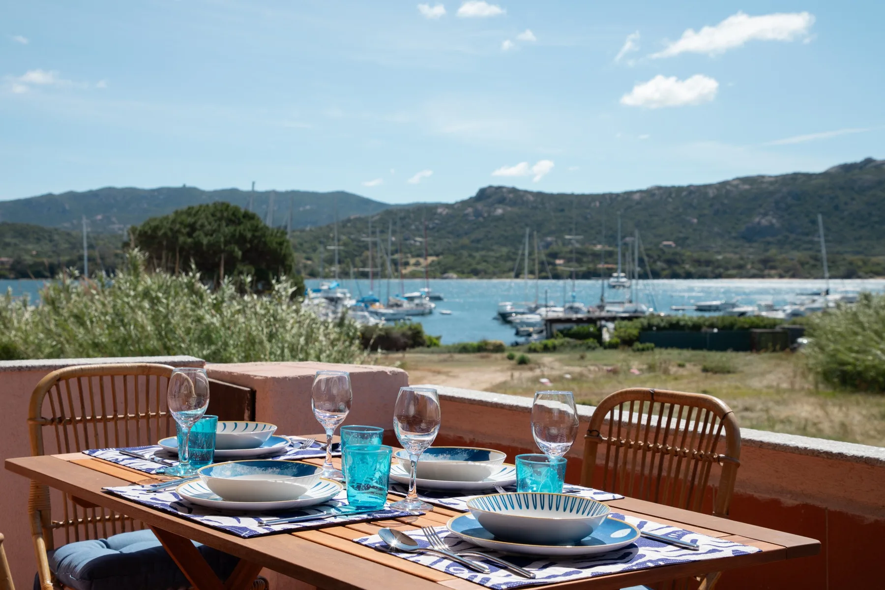 An outdoor dining table is set beside a bay with sailboats.