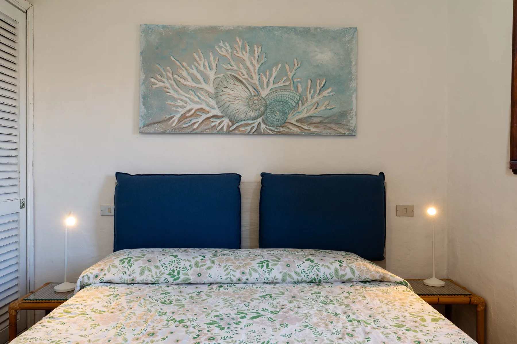 A bedroom with a bed, patterned bedding, and blue pillows beneath a coral painting.