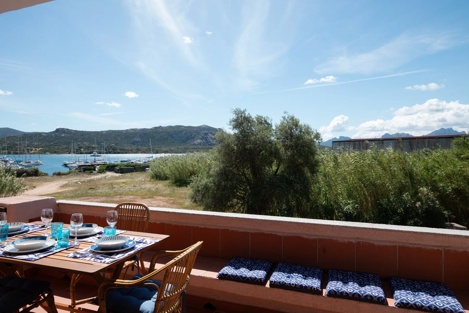 A terrace dining area overlooks shoreline vegetation, boats, and a bay.