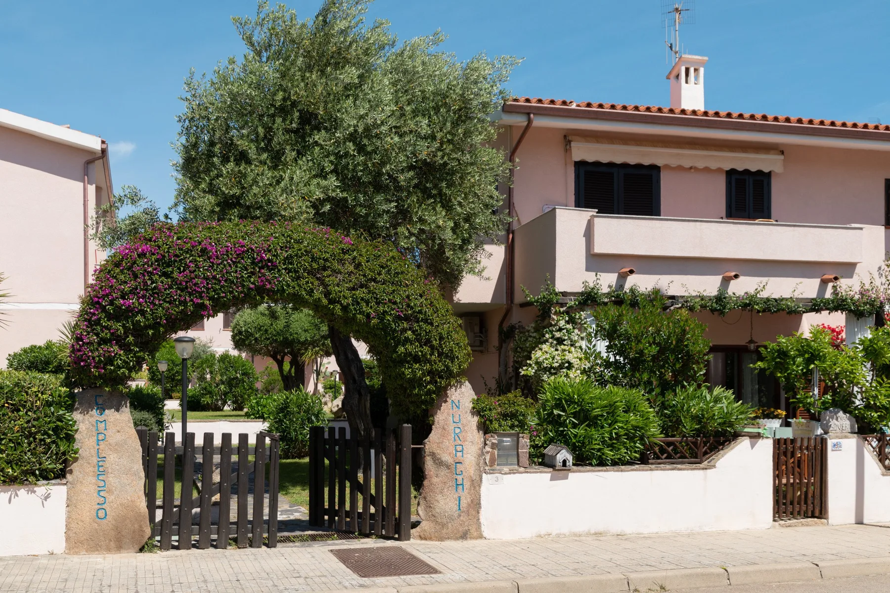 A two-story house stands behind a garden with trees and flowering shrubs.