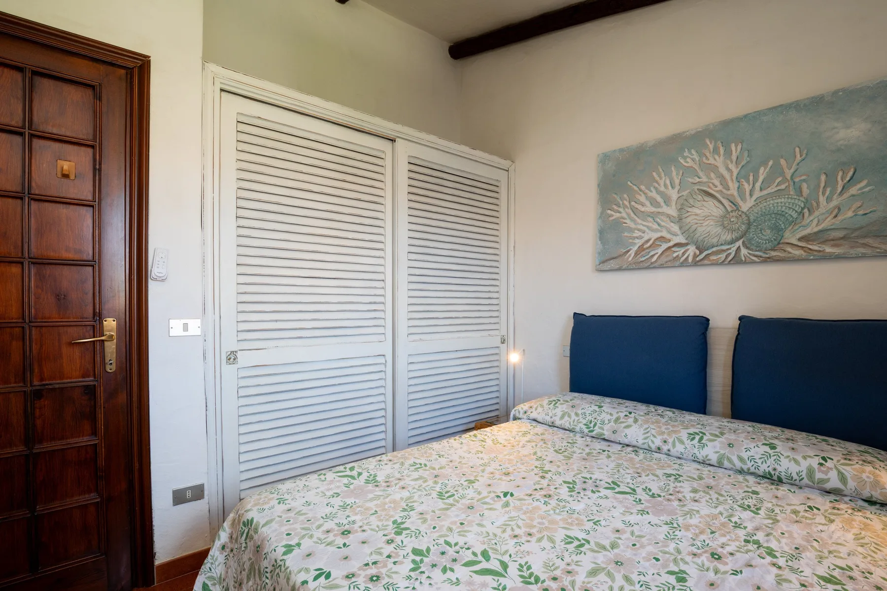 A bedroom with a floral bedspread, blue headboard, and white louvered closet doors.