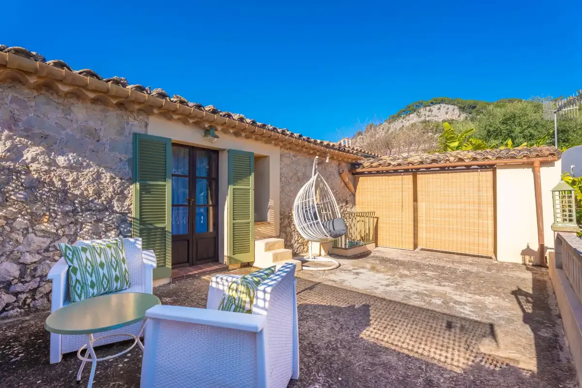 A sunlit outdoor patio has seating, a hanging chair, and shuttered doors.