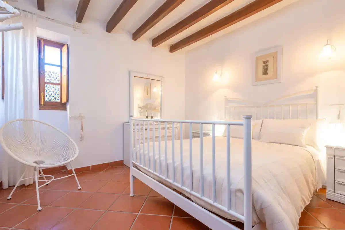 A bright bedroom with a white bed, a woven chair, and exposed ceiling beams.