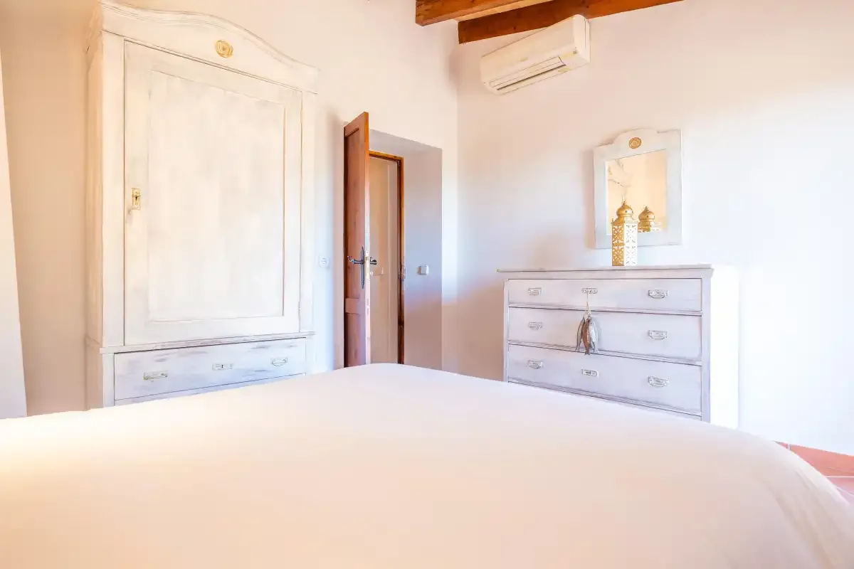 A bright bedroom with a bed, painted dressers, and exposed ceiling beams.