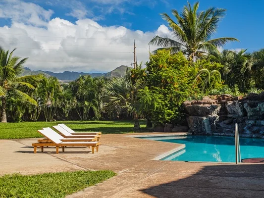 Una piscina al aire libre está junto a una terraza pavimentada, asientos reclinables y vegetación tropical.