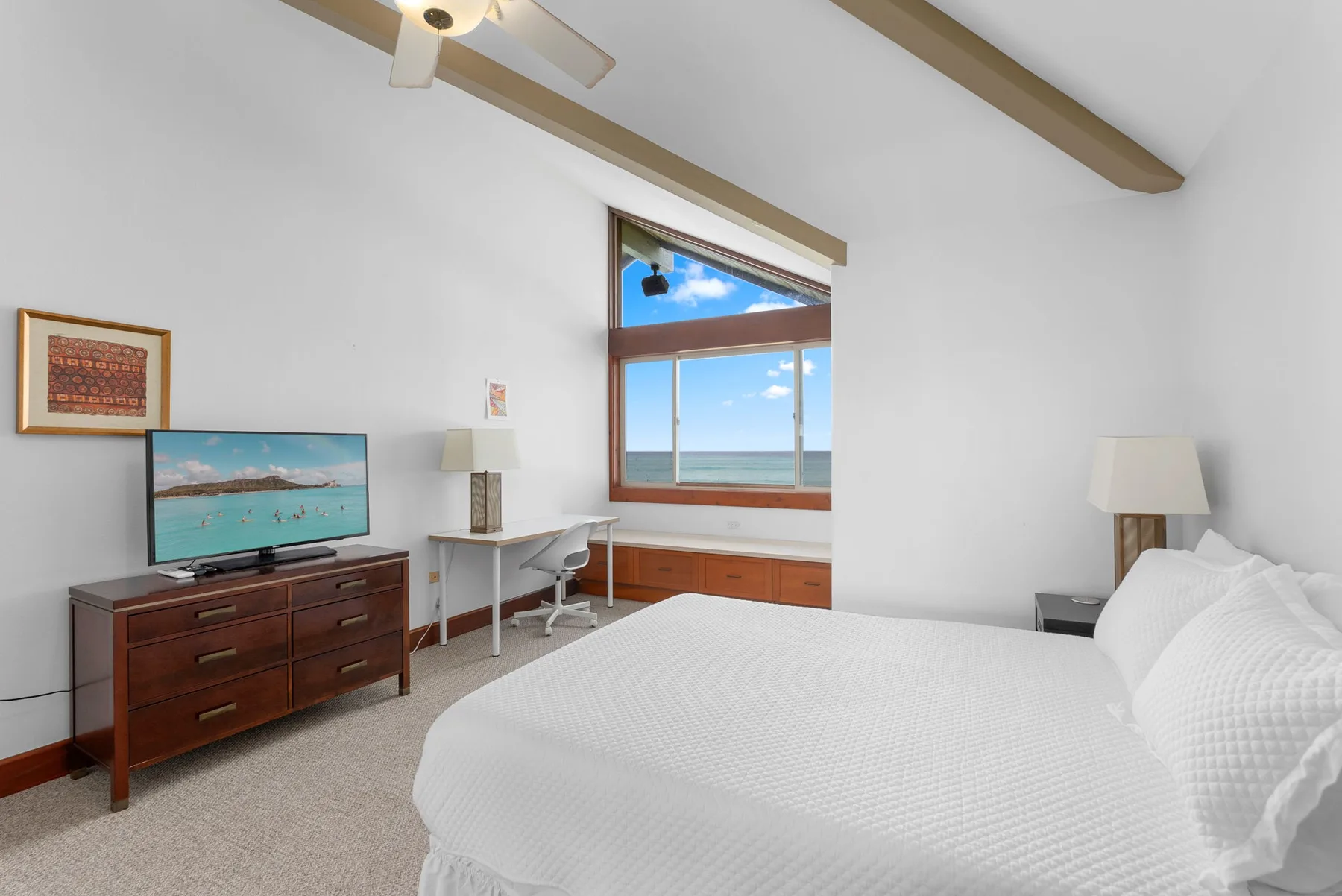 Un dormitorio luminoso con una cama grande, un escritorio y vista al mar por la ventana.