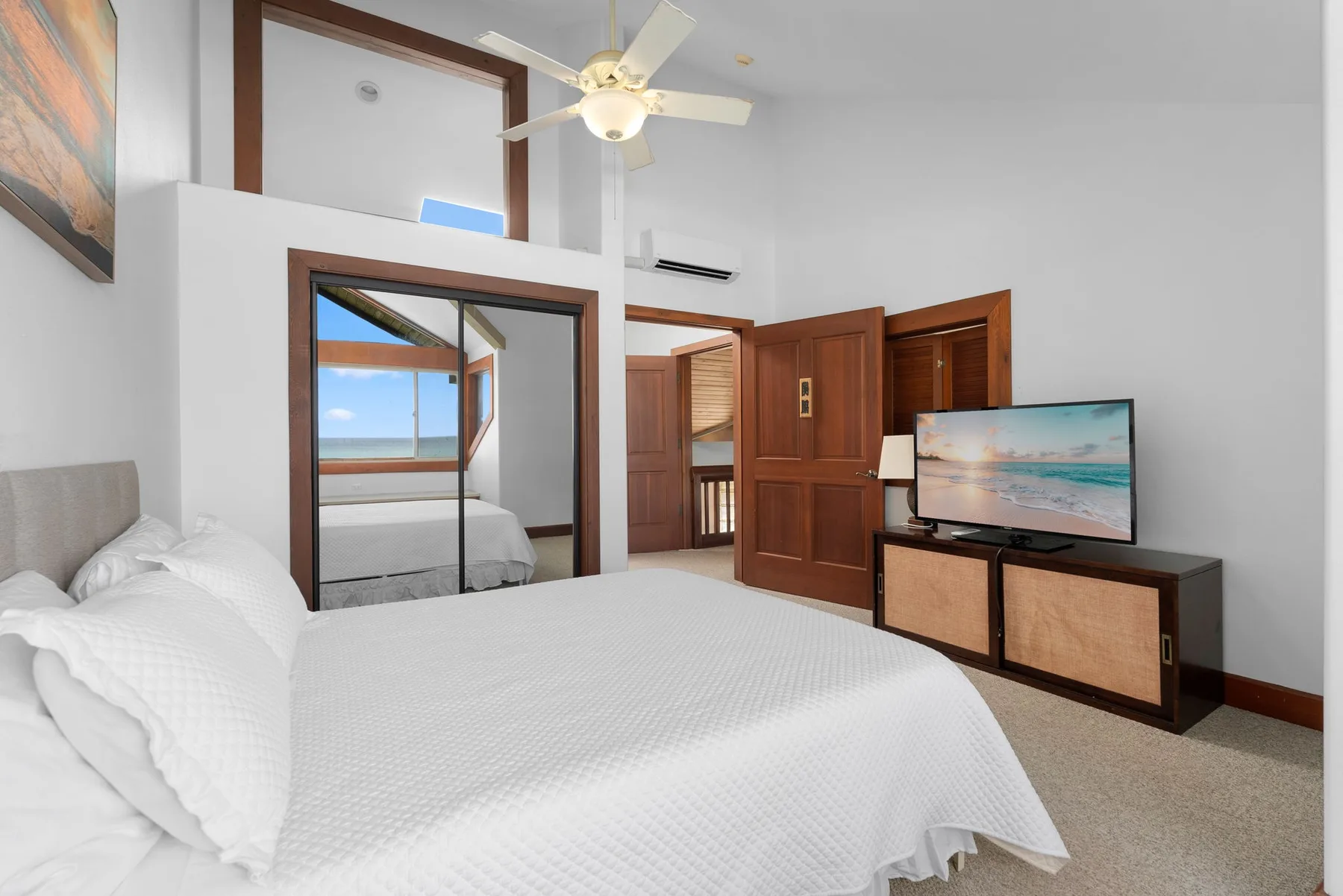 Un dormitorio luminoso con una cama blanca, puertas de armario con espejo y un televisor.