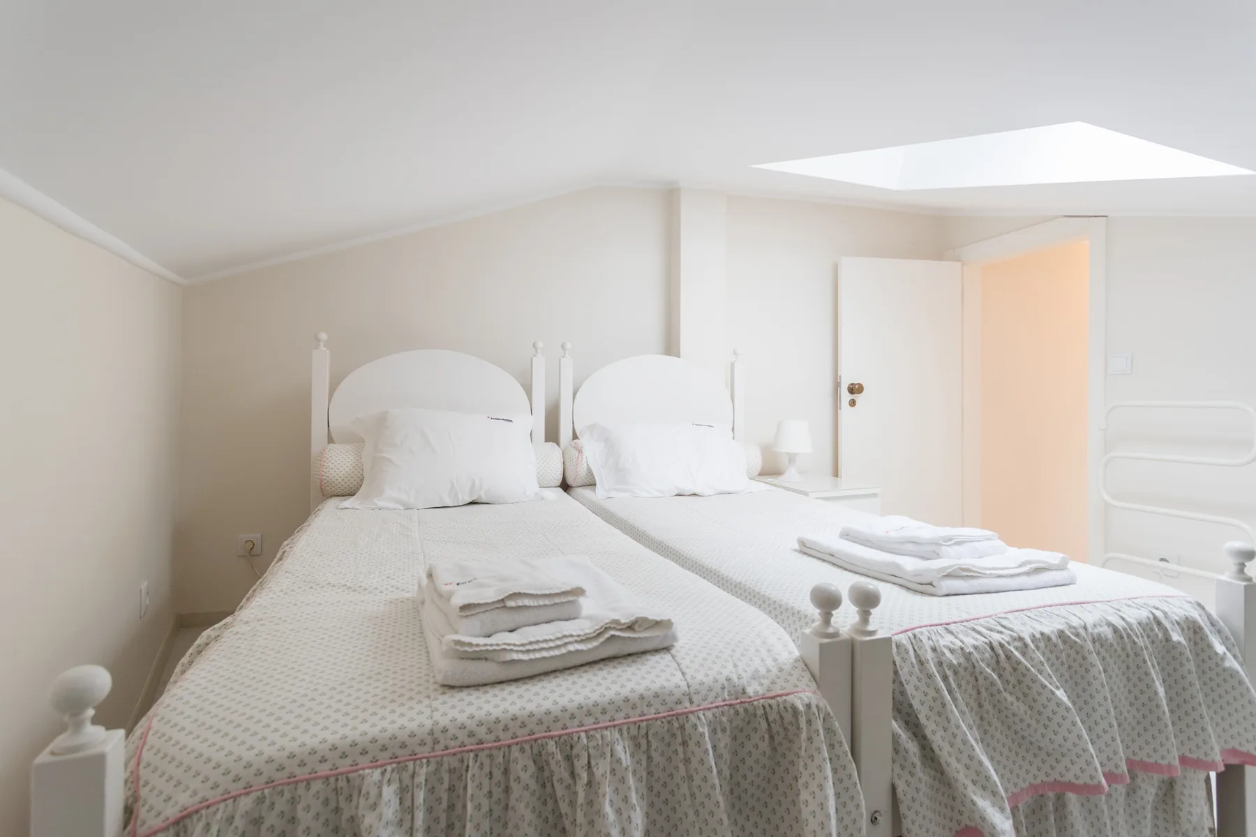 Un dormitorio luminoso con dos camas, ropa de cama blanca y una claraboya.