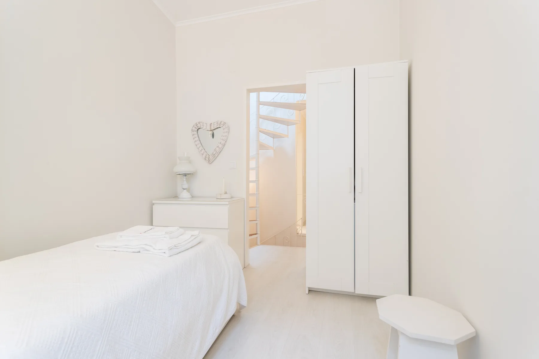 A bright bedroom with a white bed and wardrobe beside an open doorway.