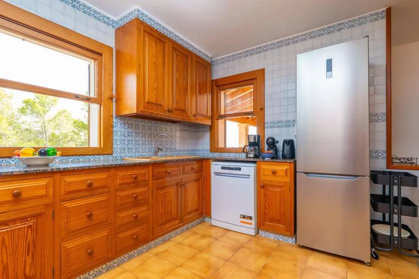 A tiled kitchen with wooden cabinets, a refrigerator, and a window overlooking greenery.