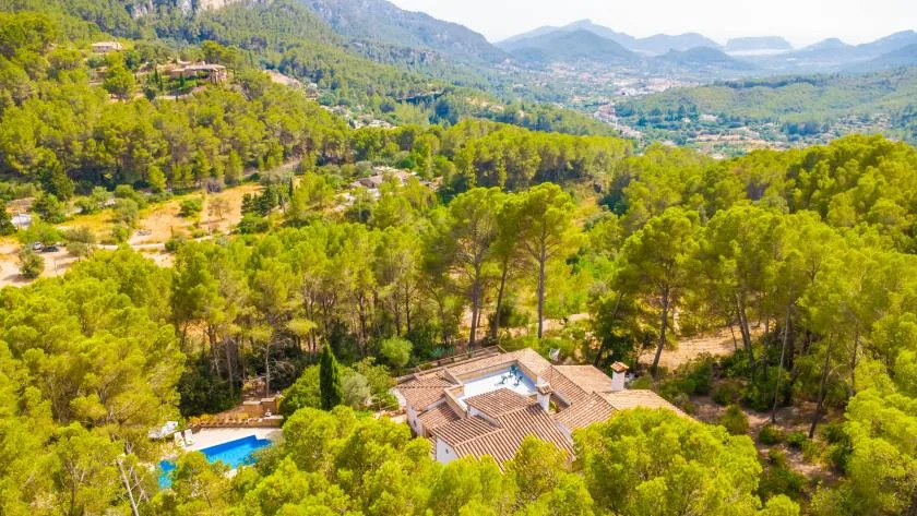 Aerial view of a tiled-roof house among wooded hills and a swimming pool.