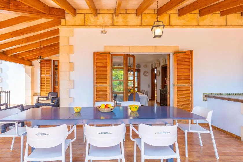 A covered dining area with a long table and wooden shutters opening to an interior room.