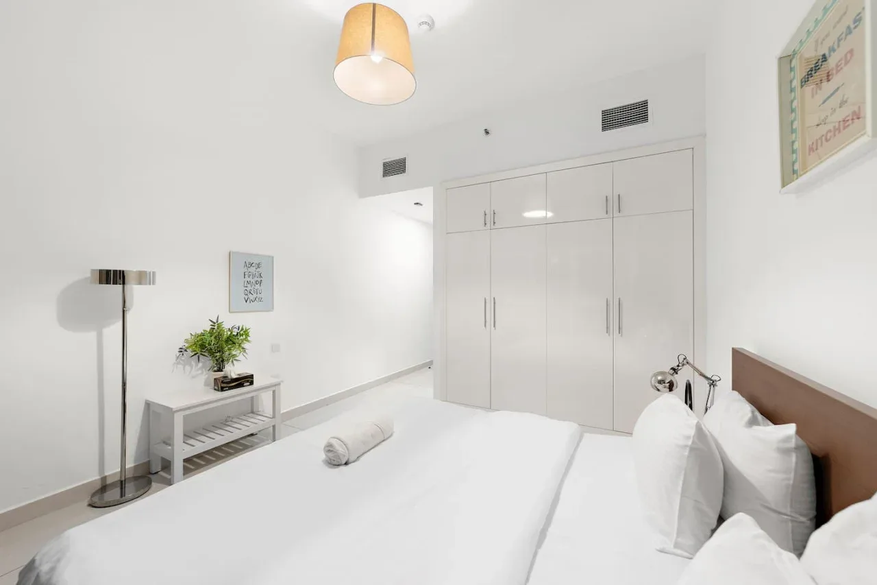 Un dormitorio luminoso con ropa de cama blanca, armarios empotrados y una lámpara.
