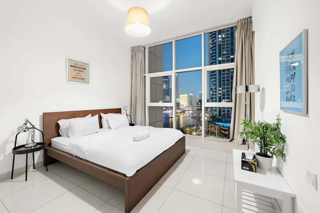 Un dormitorio luminoso con una cama tendida junto a una gran ventana con vistas a la ciudad.