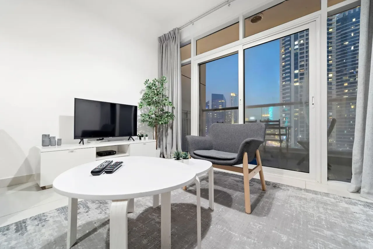 Una sala luminosa con asientos, un televisor y grandes ventanas con vistas a edificios de la ciudad.