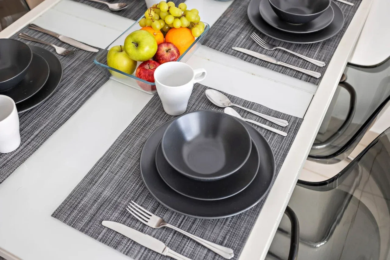Una mesa de comedor está preparada con vajilla oscura, cubiertos y un frutero.