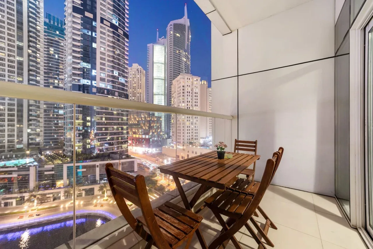 Un comedor en el balcón con asientos de madera da a edificios altos iluminados.