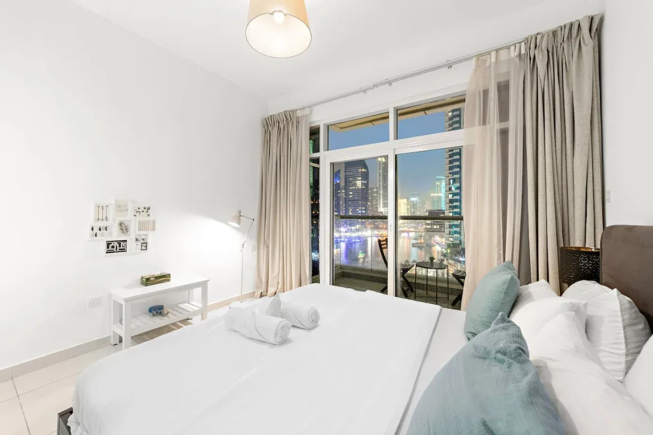 Un dormitorio luminoso con una cama junto a grandes ventanas con vistas a edificios de la ciudad.