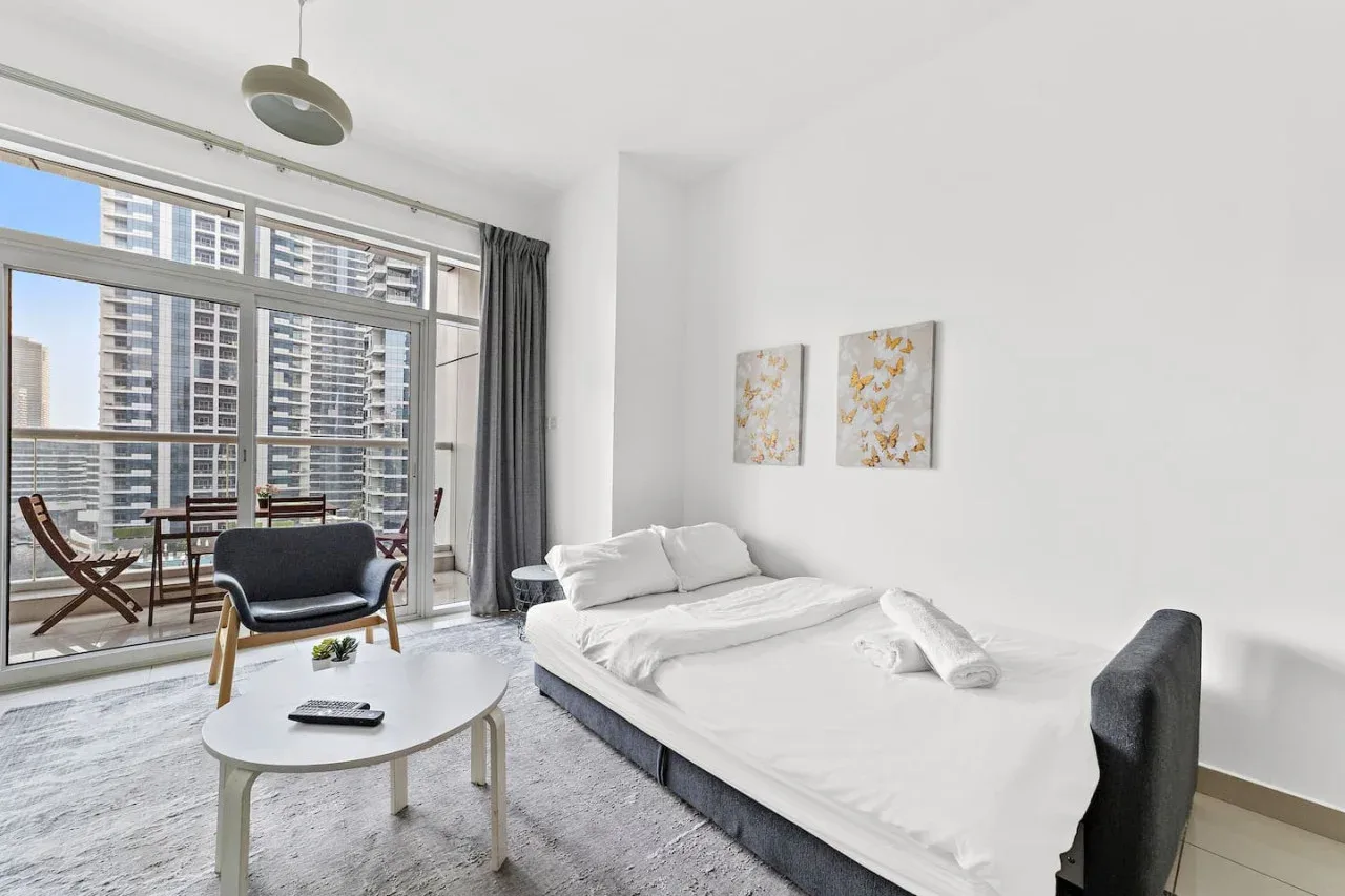 Un dormitorio luminoso con una cama junto a grandes ventanales con vistas a edificios cercanos.