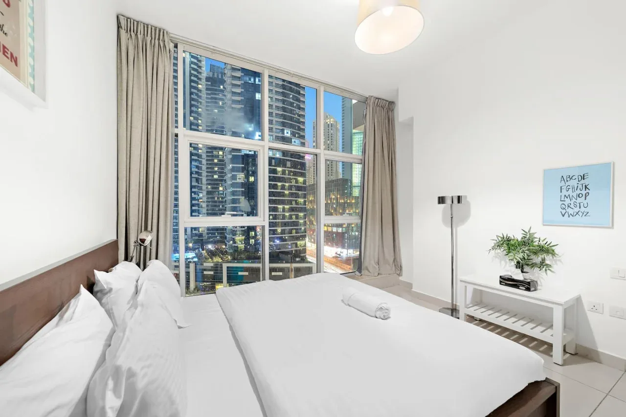 Un dormitorio luminoso con una cama grande y ventanales que dan a edificios cercanos.