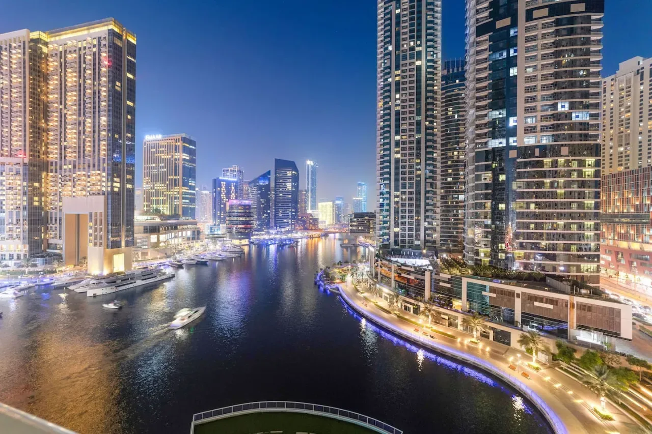 Un paseo marítimo urbano de noche, con edificios altos iluminados y barcos en el agua.