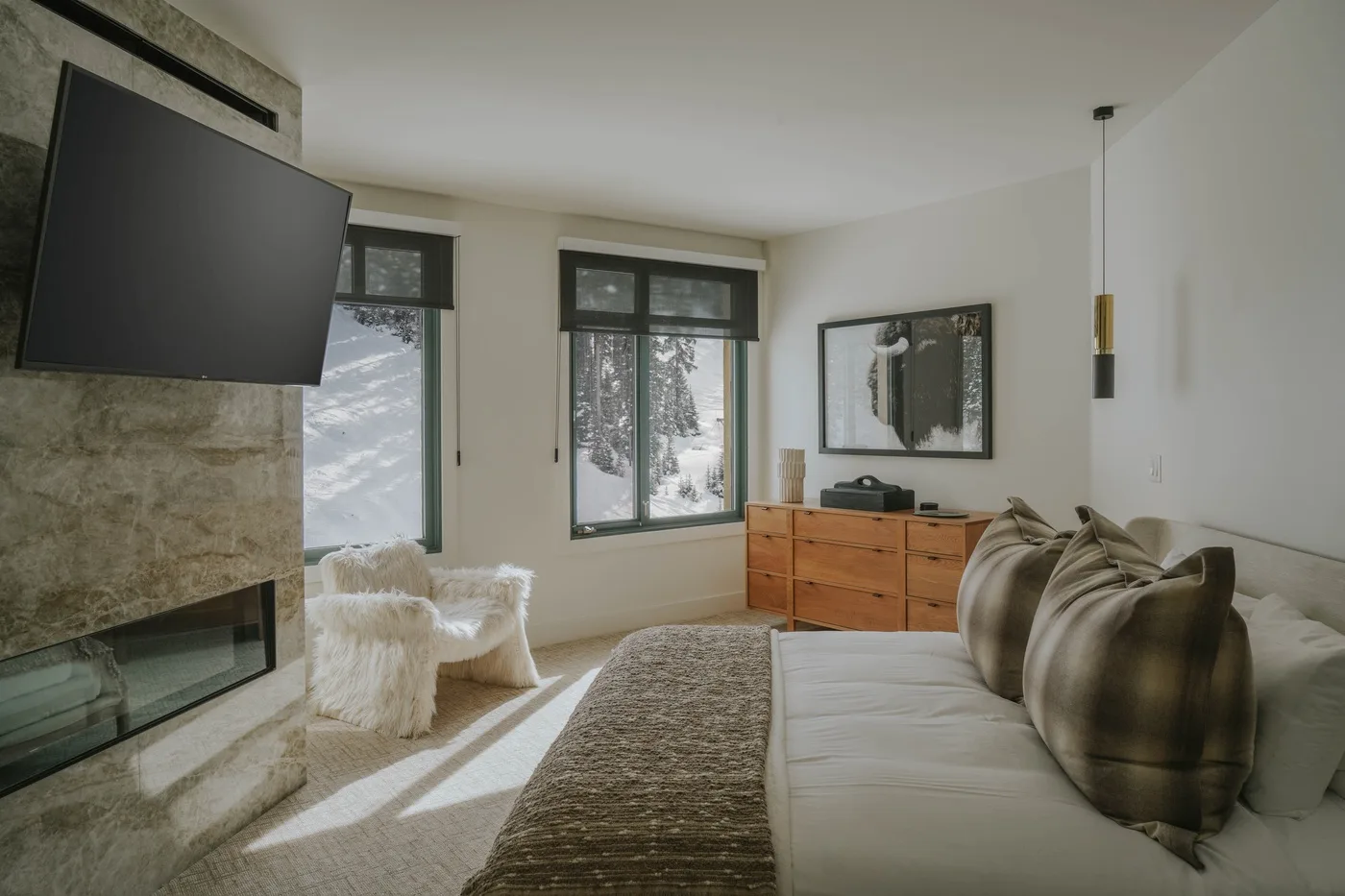 A bright bedroom with a bed, stone fireplace, and windows overlooking snowy trees.