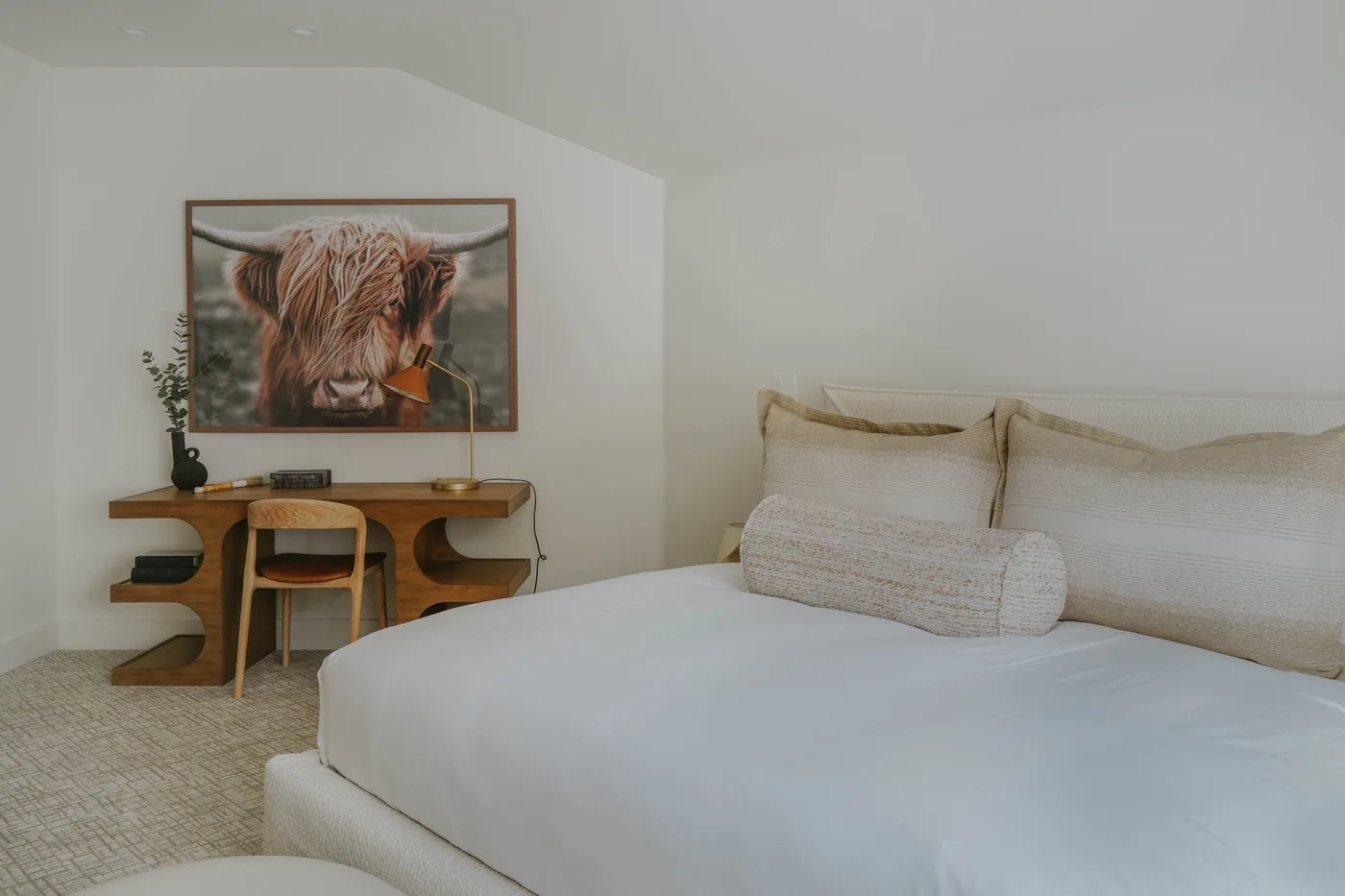 A bright bedroom with a large bed and a wooden desk beneath framed artwork.