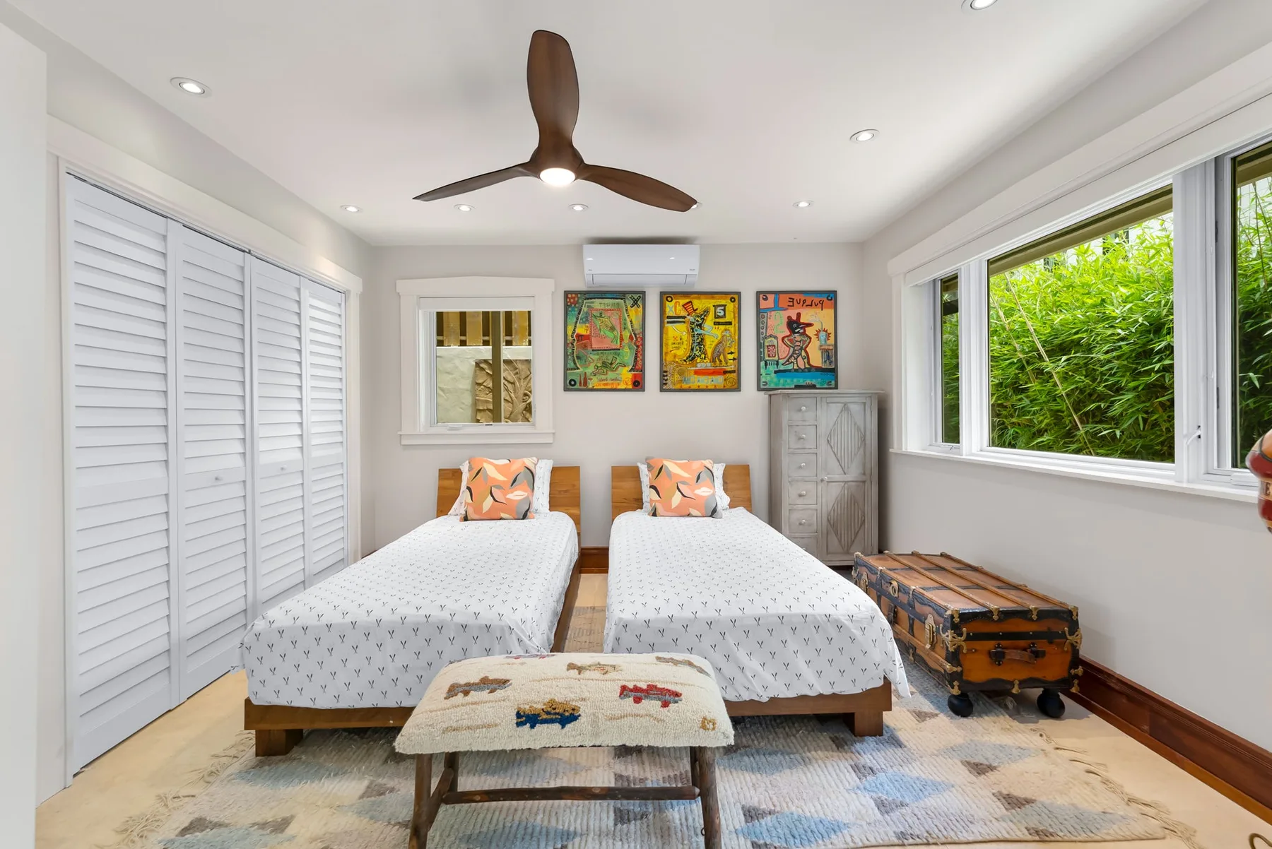 Un dormitorio luminoso con dos camas, arte colorido en la pared y grandes ventanas con vistas a la vegetación.