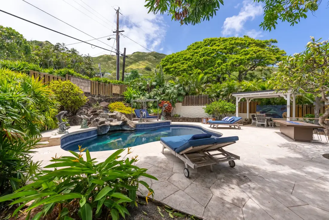 Una zona de piscina al aire libre con tumbonas, vegetación y un patio cubierto.