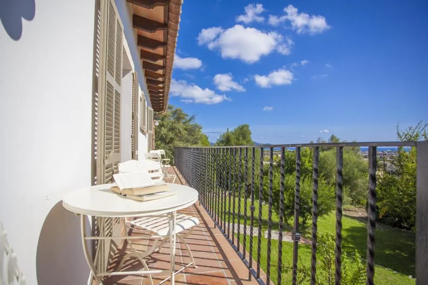 A sunny balcony with outdoor seating overlooks green trees beneath a blue sky.