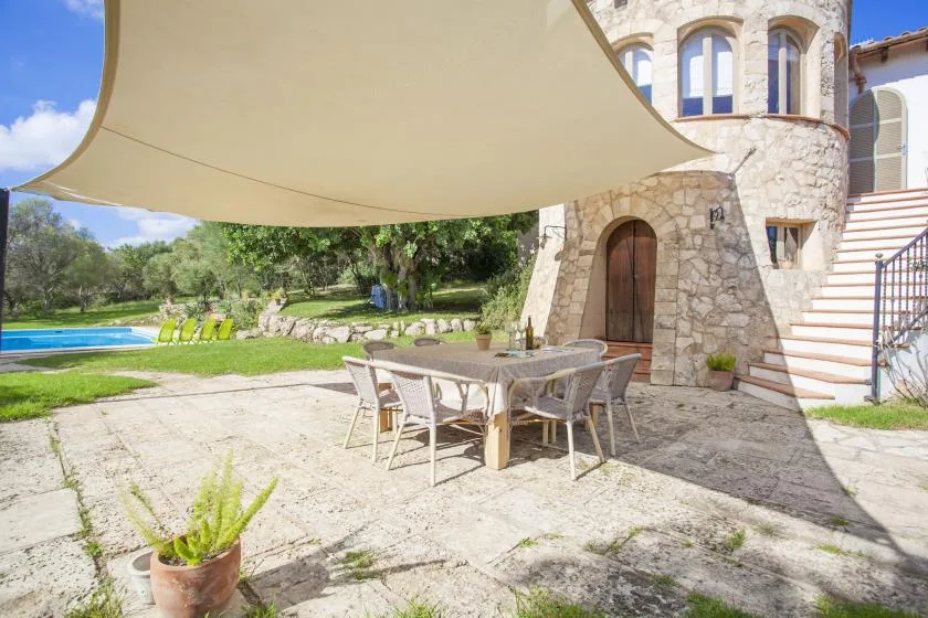 A shaded outdoor dining area beside a stone building, with a pool and garden beyond.