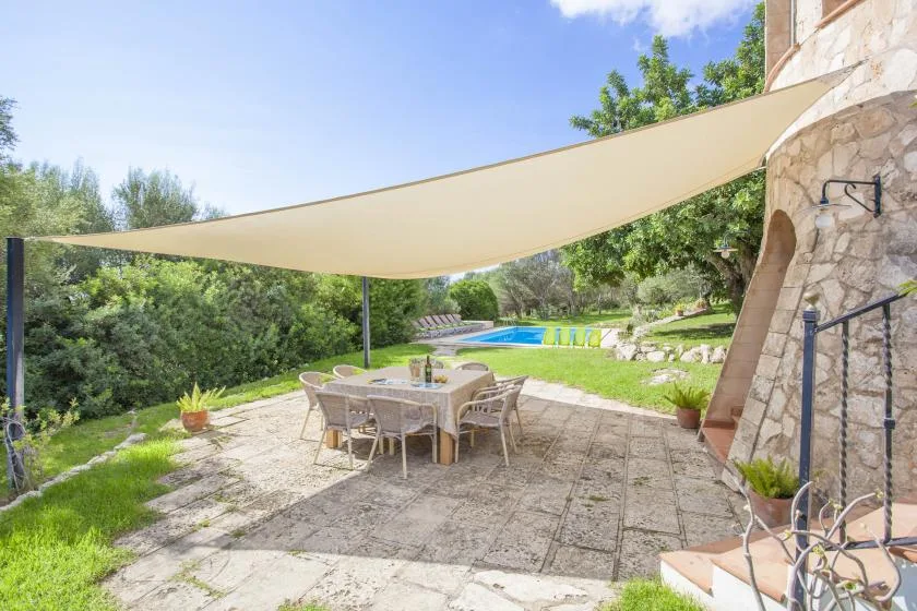 An outdoor patio dining area beneath a shade canopy, overlooking a swimming pool and garden.