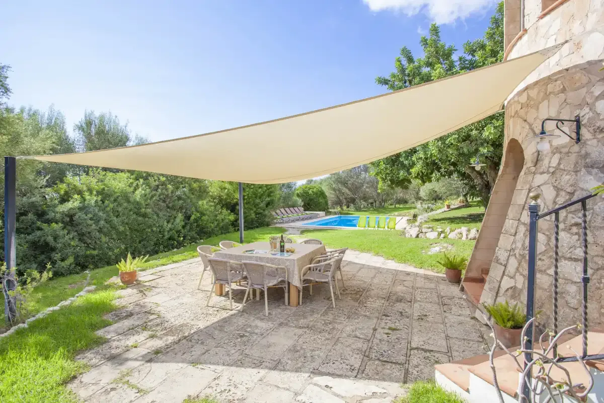 A shaded outdoor patio with a dining table overlooks a swimming pool and garden.