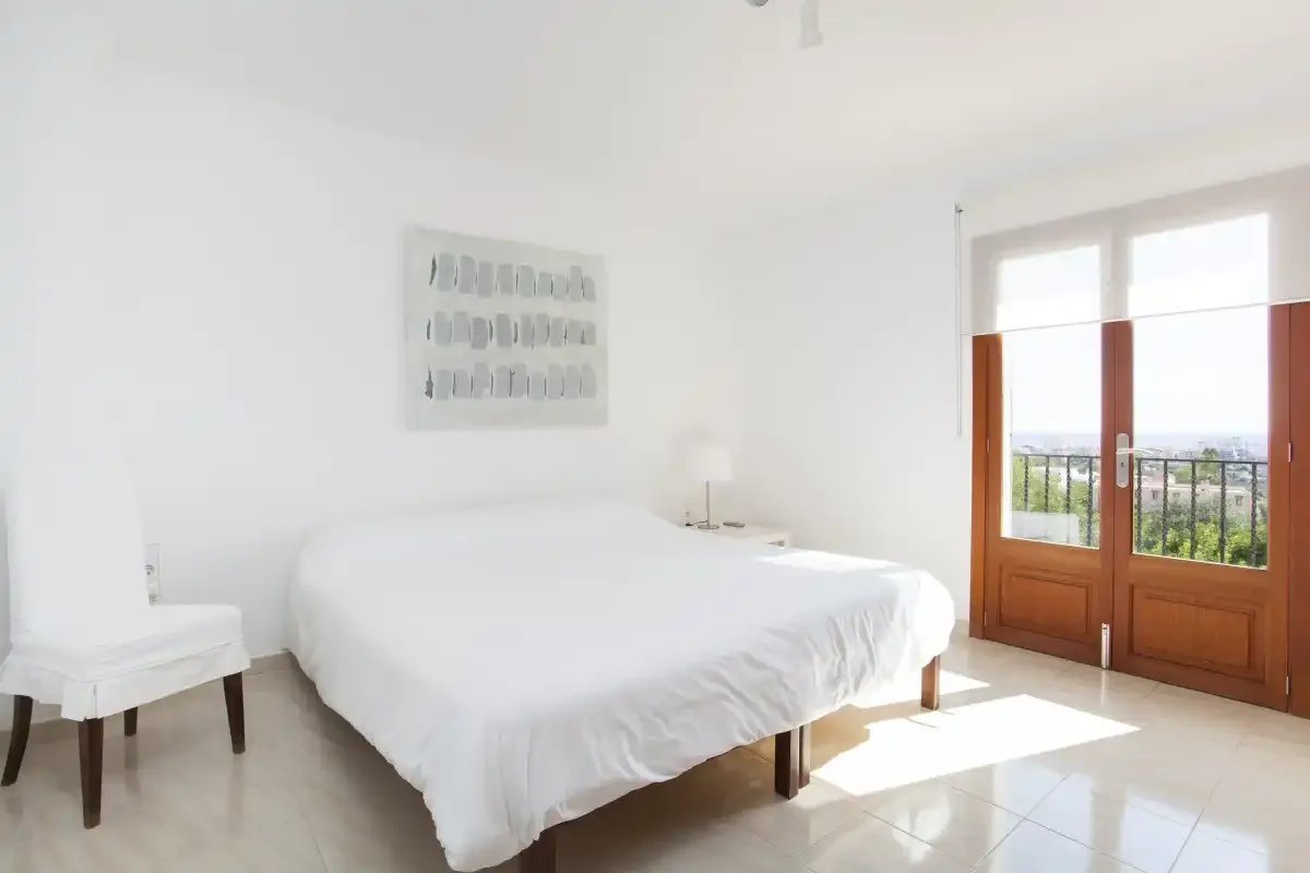 A bright bedroom with a white bed beside glass doors opening onto a balcony.