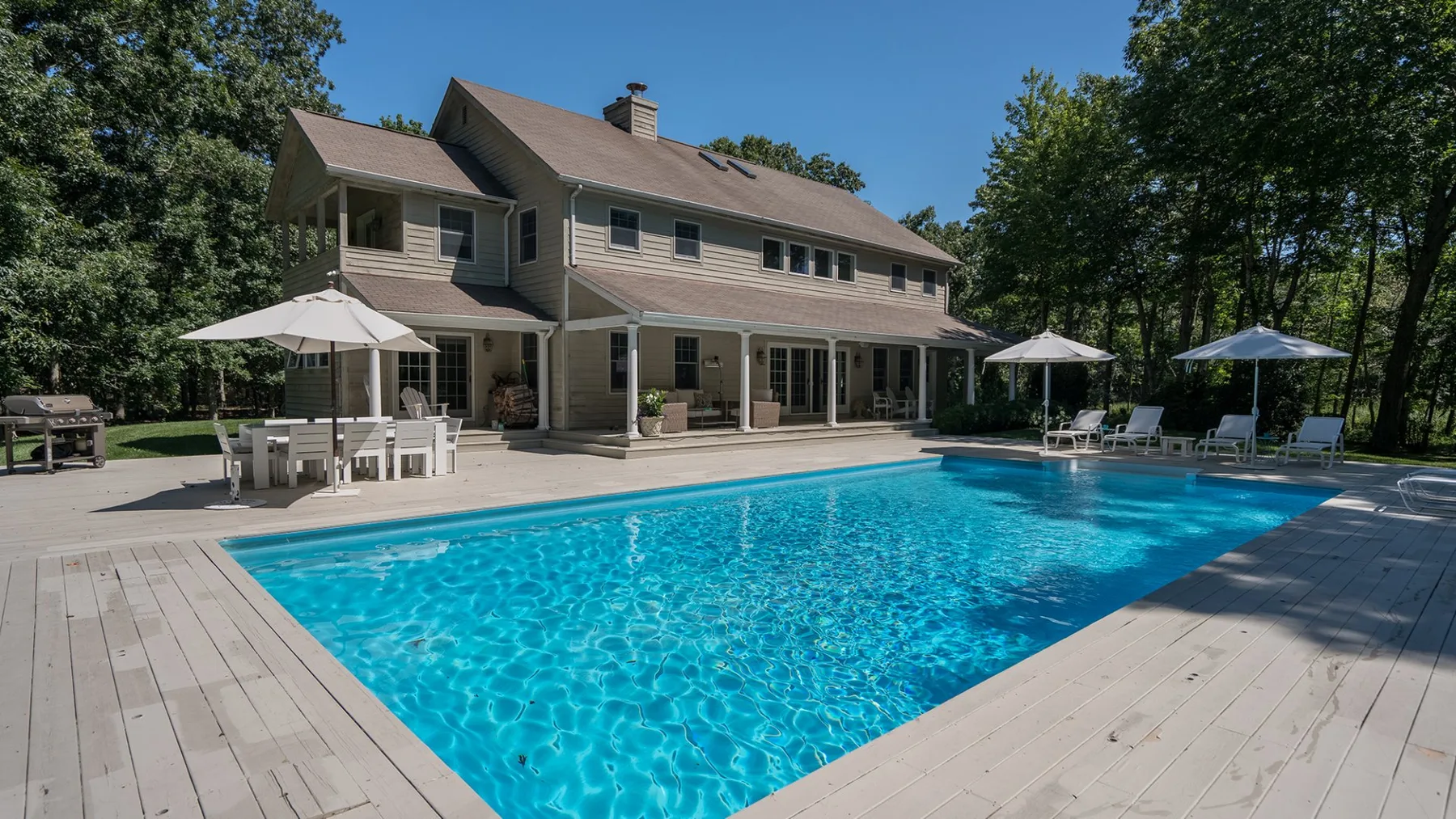 A swimming pool beside a large house, with patio seating and umbrellas.
