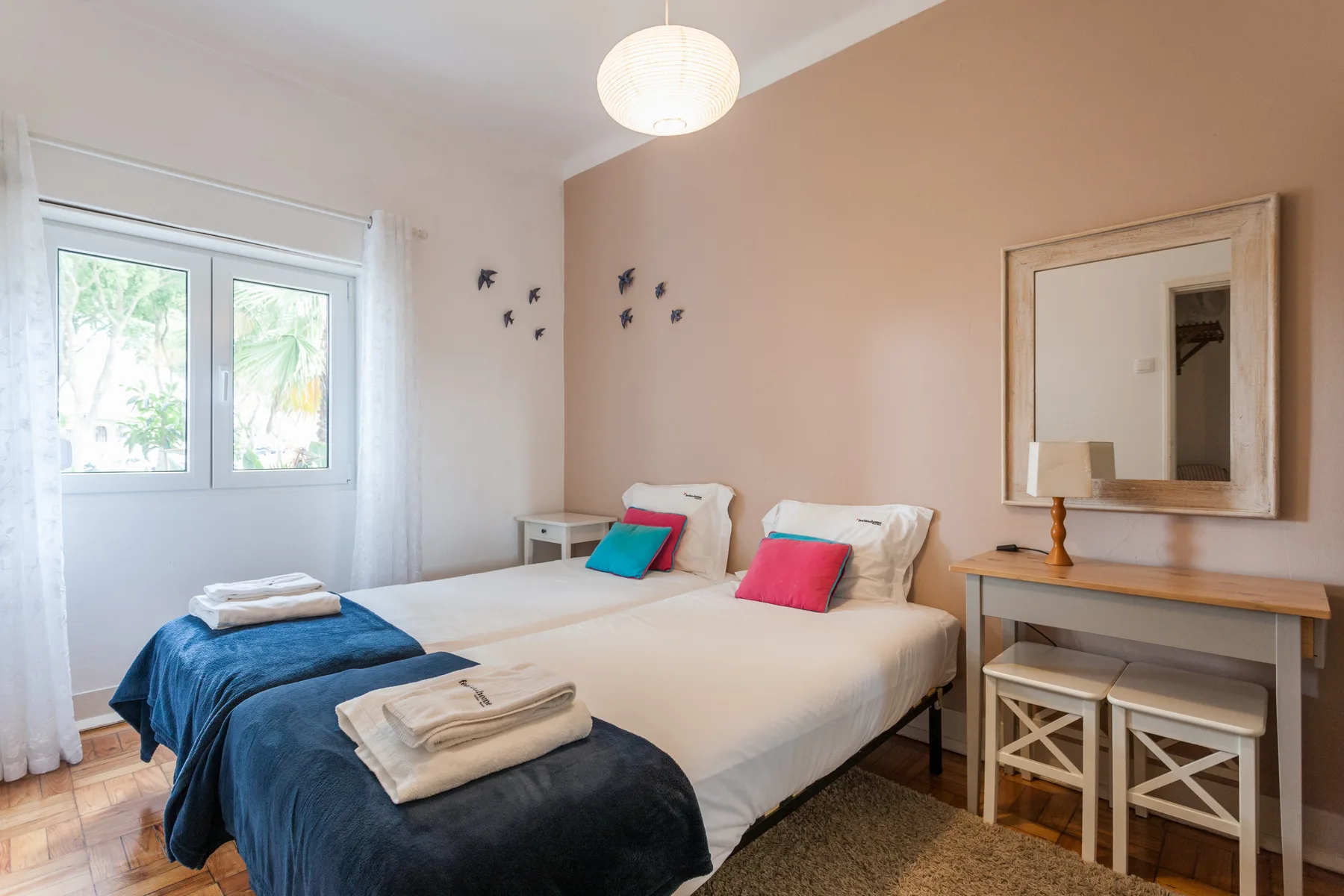 Un dormitorio luminoso con dos camas, cojines de colores y una ventana.