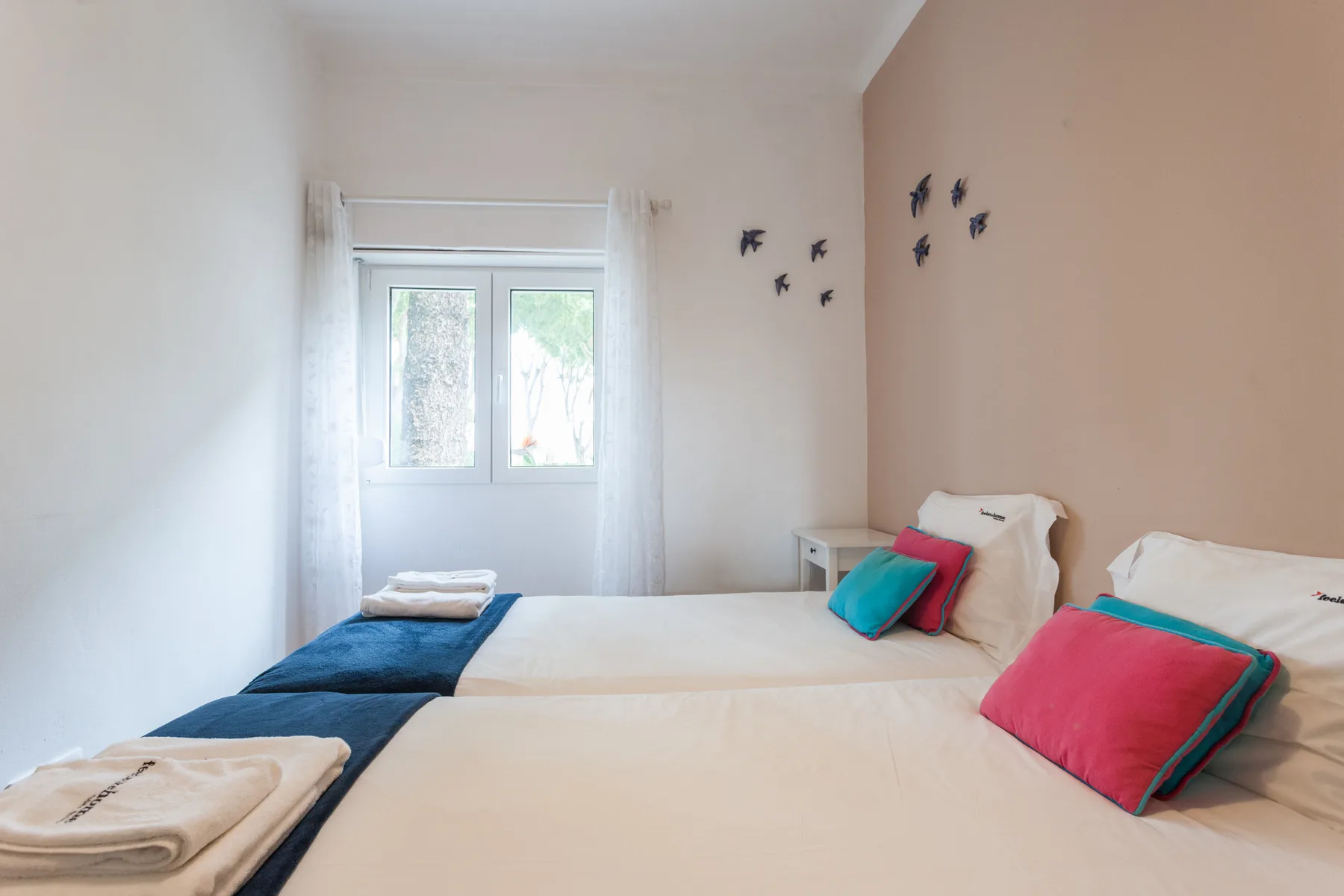 Un dormitorio luminoso con dos camas, cojines de colores y una ventana.